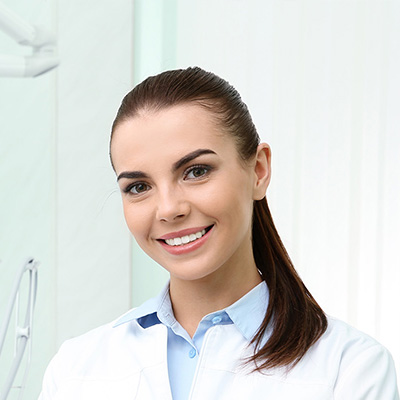 A smiling woman with brown hair, wearing a white coat and blue shirt, stands in front of a dental office setting.